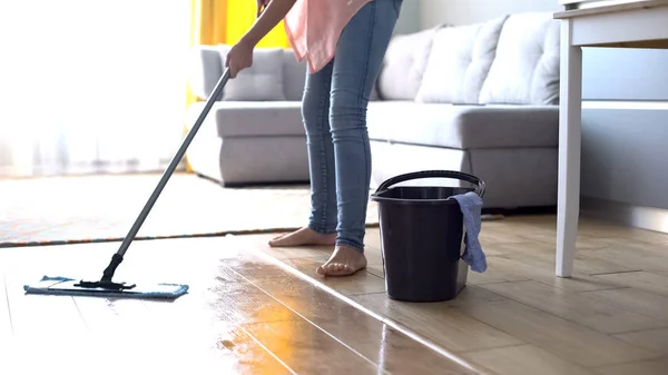 Person mopping wooden floor near gray sofa and bucket